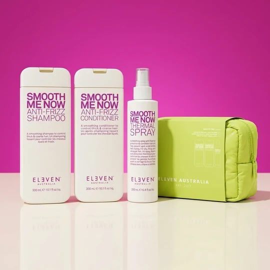 A haircare set from ELEVEN AUSTRALIA including Smooth Me Now Anti-Frizz Shampoo, Smooth Me Now Anti-Frizz Conditioner, Smooth Me Now Thermal Spray, and a lime green zippered pouch, arranged on a reflective white surface against a vibrant magenta-pink background.
