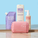 A set of Eleven Australia hair color care products arranged on a reflective surface against a light blue background, including a pink zip pouch, a purple treatment pouch, a white shampoo bottle, and a beige hair spray bottle.