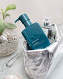A teal pump bottle labeled KEPLMURPHY FLAWLESS hair product sits in a shiny silver pouch beside a glass bottle, with a small plant and pale stones in the background - Hair Care Canada