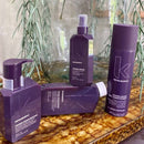 Four Kevin Murphy Young Again haircare products in a coordinated deep purple color, arranged on a polished brown marble surface with a glass vase and green leaves in the background.