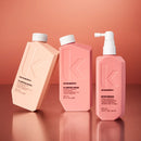 Three Kevin Murphy haircare bottles in a studio setup: a peach-colored Plumping Wash bottle, a pink Plumping Rinse bottle, and a pink Body Mass pump bottle, arranged on a reflective peach surface against a peach background.