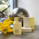 Kevin Murphy Smooth Again hair care set including a pale yellow pump bottle, a white box, and a yellow box, styled on a stone surface with a yellow lily flower and a metallic sphere in the background.