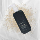 Kevin Murphy Thock-Again thickening agent in a black bottle with a white pump dispenser, styled on a white fabric background with gold glitter.